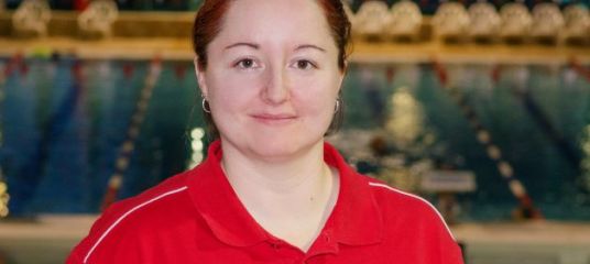 Landestrainerin Christina Bauer im roten T-Shirt. Im Hintergrund ein Schwimmbecken.